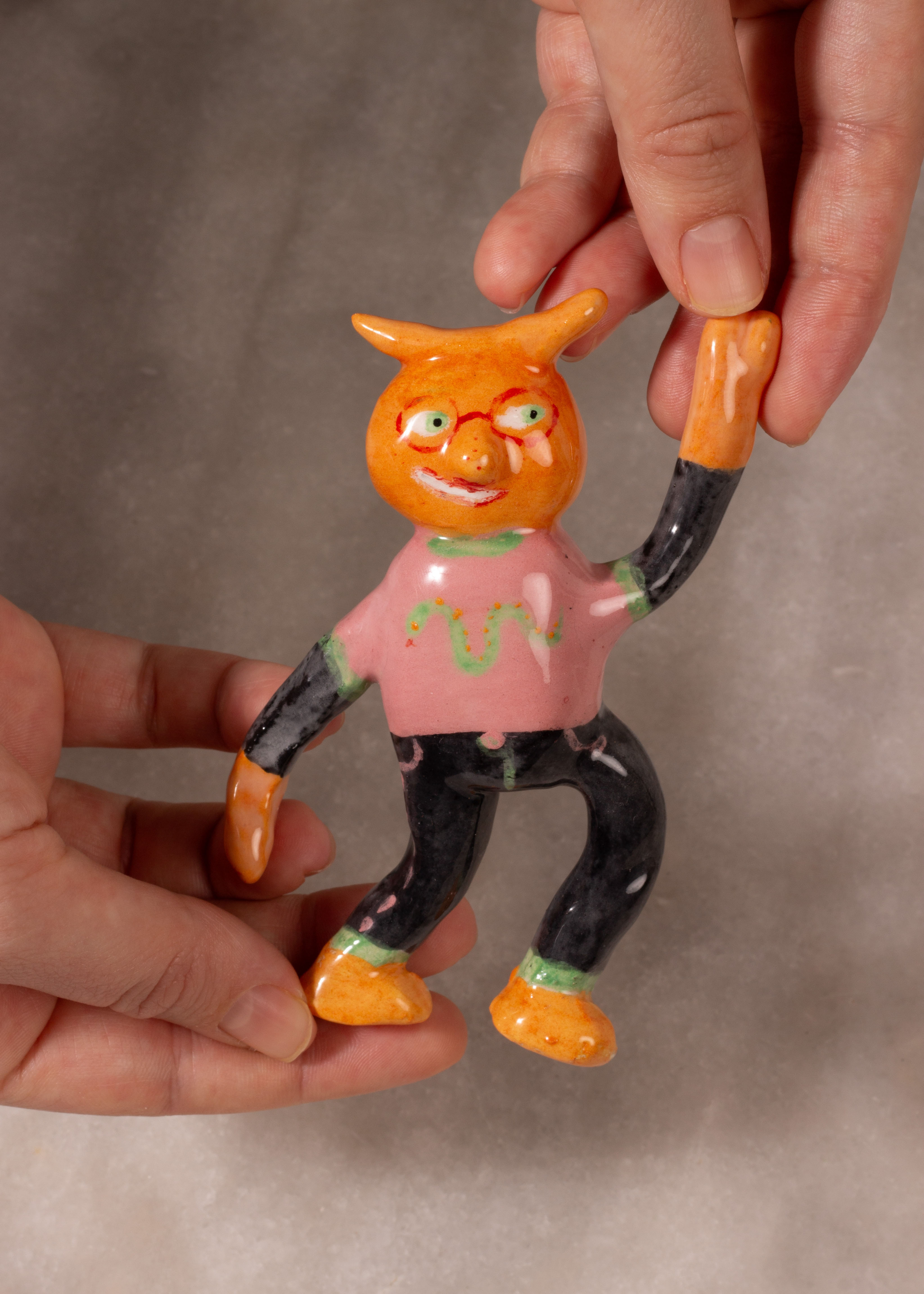 petit personnage orange souriant en faïence avec des lunettes rouges, des cornes, un jean noir et un t shirt rose avec un serpent vert