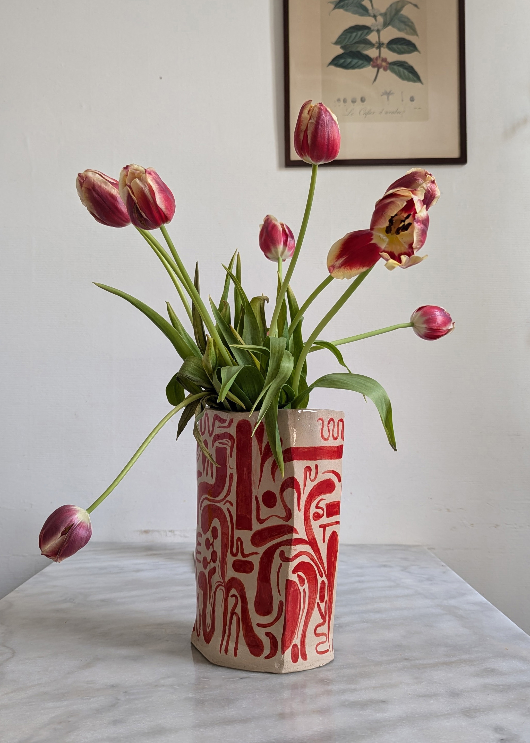 grand vase en faïence blanche peinte avec des motifs abstraits rouges, dans lequel on voit un bouquet de tulipes rouges et jaunes, posé sur une table en marbre